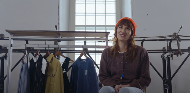 Person sitting in front of a track sewing machine, next to a clothing rack with a few articles of clothing on it.
