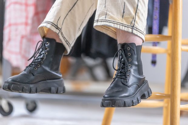 A pair of feet hanging down from a wooden chair. They have black lace-up boots on, and the bottoms of their pants are visible. The pants are off-white and detailed with black to give it a 2D effect.