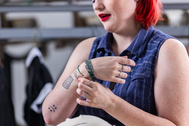 Person holding their arm up, gesturing to a handwritten tattoo. They have on beaded bracelets and silver rings, and two more visible tattoos on their arm.
