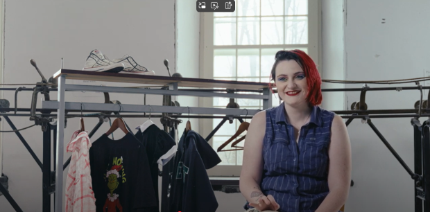 Person sitting in front of a track sewing machine, next to a clothing rack with a few articles of clothing on it.