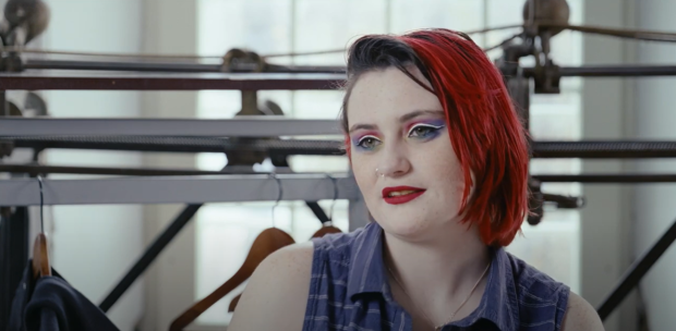Close-up of a person in front of a track sewing machine and clothing rack, in interview format.