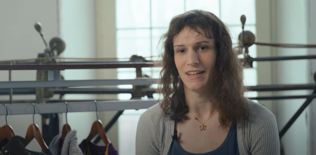 Close-up of a person in front of a track sewing machine and clothing rack, in interview format.