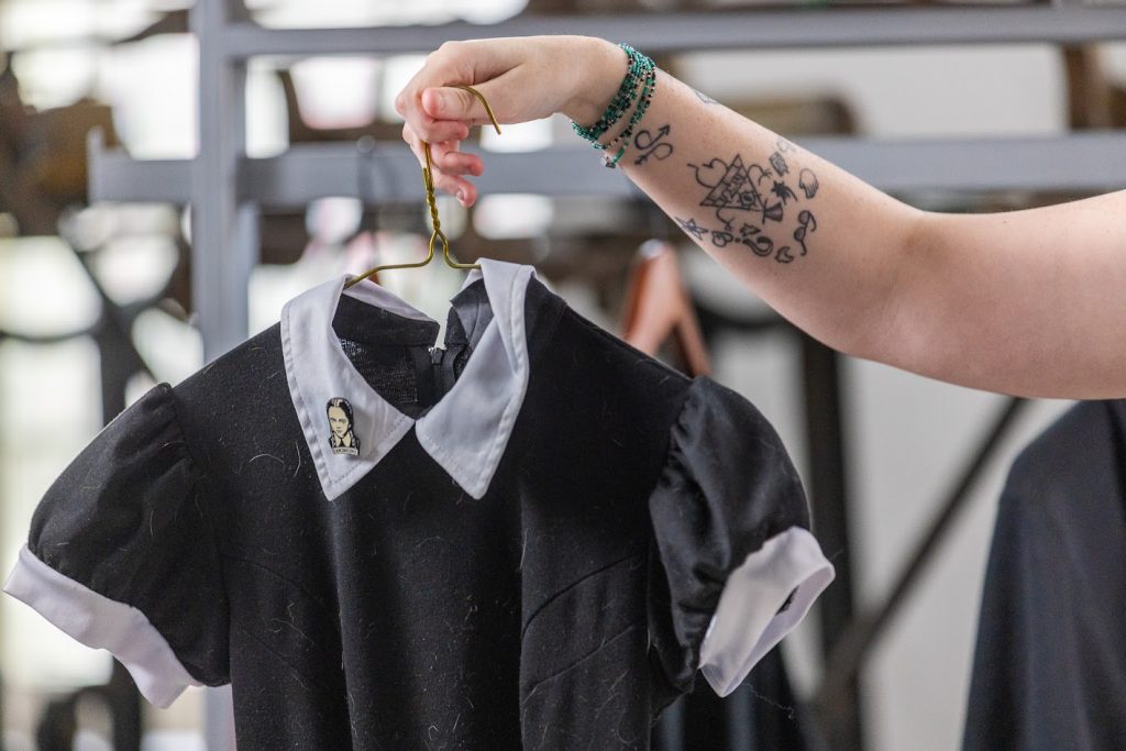 An arm with several small, black tattoos is holding a metal hanger. A black dress with a white collar and white cuffs around the short, puff sleeves, is on the hanger. A black and white enamel pin of a girl with two braids, Wednesday Addams, is pinned to the left side of the collar.