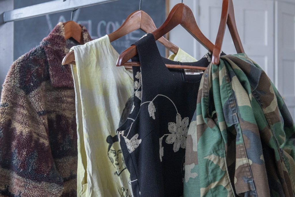 Four articles of clothing lined up in a row, hanging on wooden hangers, showing a range of style. There is a fuzzy cardigan with a landscape pattern, tank top with a black graphic, black sleeveless dress with a white floral pattern, and camo pants.
