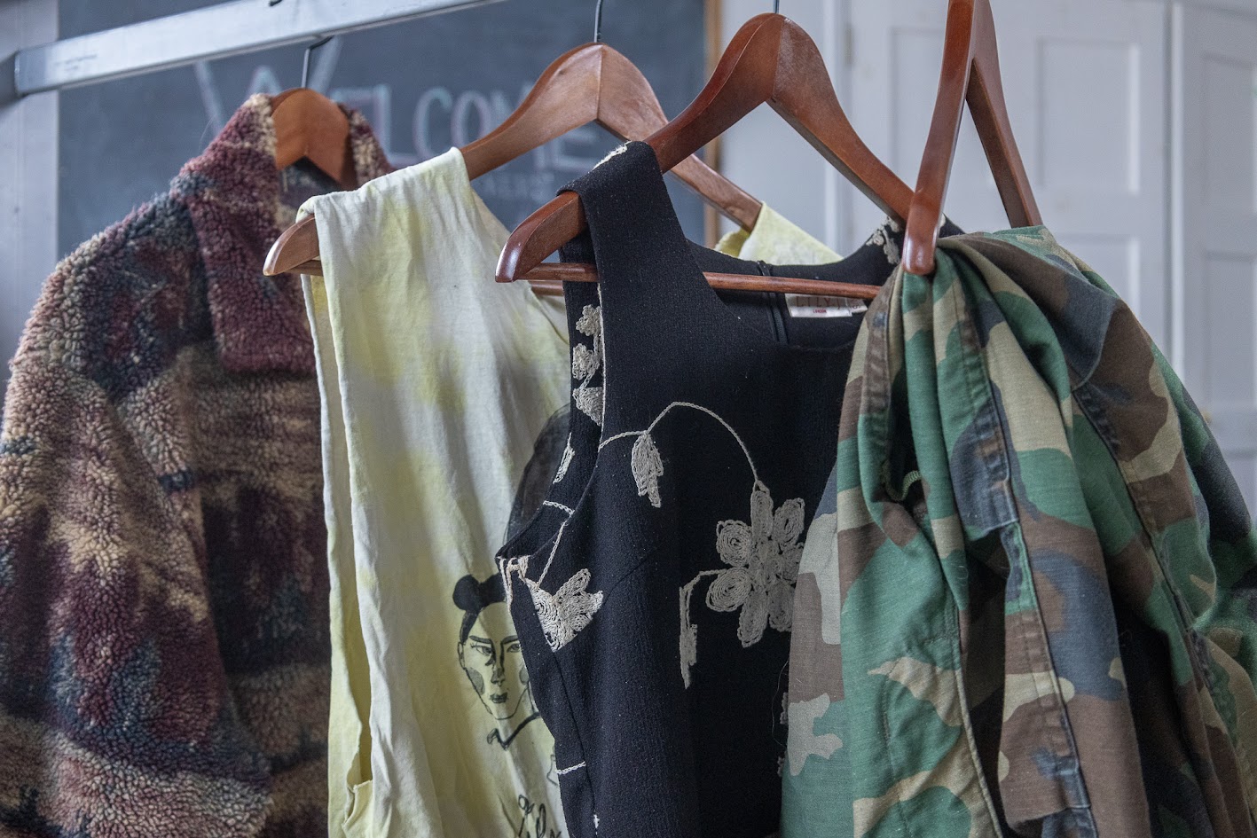 Four articles of clothing lined up in a row, hanging on wooden hangers, showing a range of style. There is a fuzzy cardigan with a landscape pattern, tank top with a black graphic, black sleeveless dress with a white floral pattern, and camo pants.