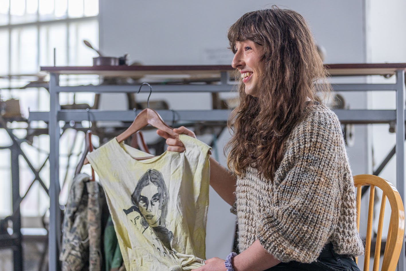 Person holding up a tank top on a wooden hanger. The top is white and yellow tie-dye, with a black graphic of faces.