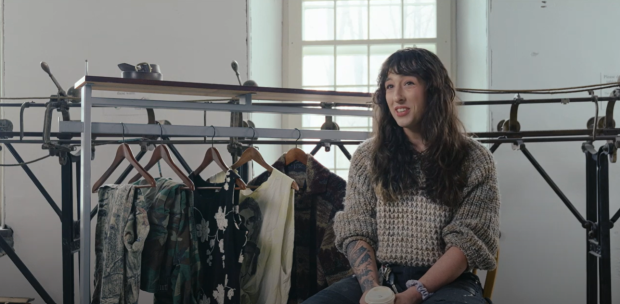 Person sitting in front of a track sewing machine, next to a clothing rack with a few articles of clothing on it.