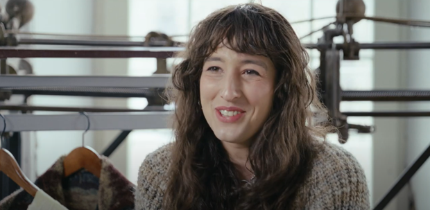 Close-up of someone in front of a track sewing machine and clothing rack, in interview format.