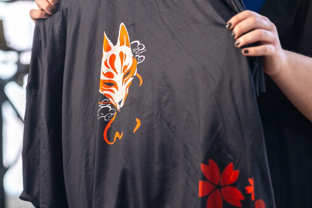 A pair of hands holding up a black shirt with a white, orange, and red graphic of a kitsune face (fox spirit), and red flowers.