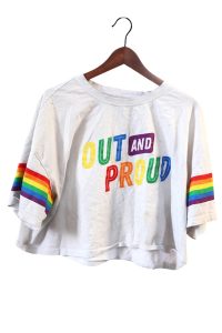An off-white cropped top on a wooden hanger. Rainbow-coloured text reads OUT AND PROUD. Rainbow-coloured stripes wrap around the sleeves.