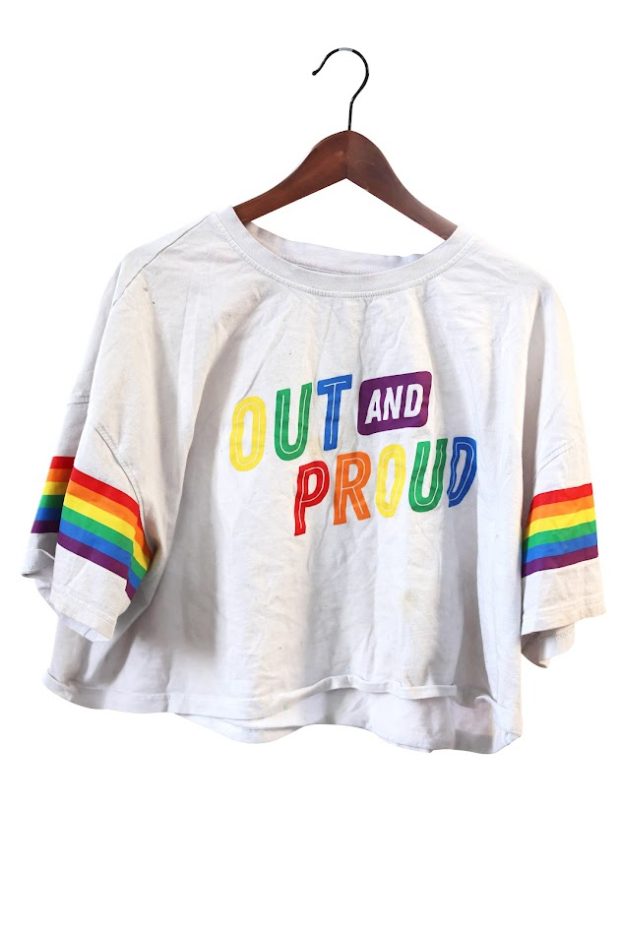An off-white cropped top on a wooden hanger. Rainbow-coloured text reads OUT AND PROUD. Rainbow-coloured stripes wrap around the sleeves.