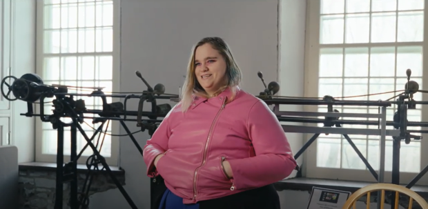 Person standing in front of a track sewing machine, wearing a bright pink jacket.