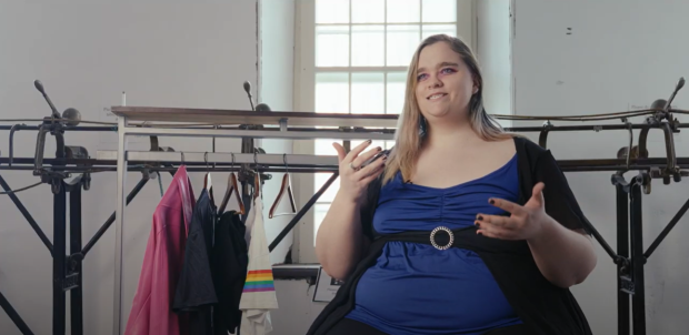 Person sitting in front of a track sewing machine, next to a clothing rack with a few articles of clothing on it.