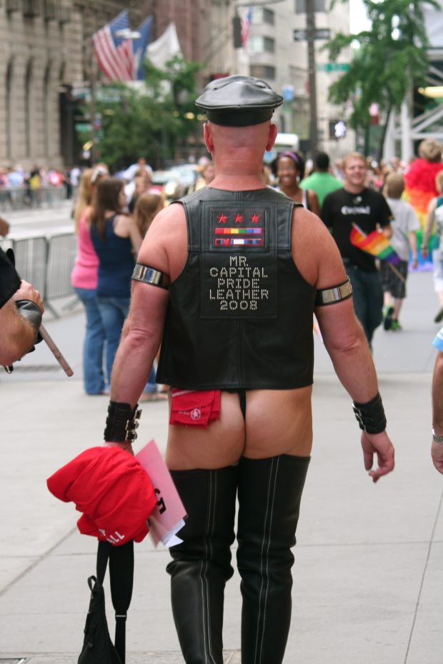 Back of a man standing outside on a busy sidewalk. He is wearing thigh-high leather boots, a thong with a red handkerchief tucked into the band, a leather cap, and a leather vest with bedazzled text reading MR CAPITAL PRIDE LEATHER 2008.