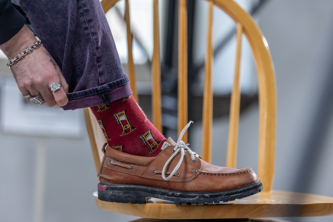 A foot is resting on a light wooden chair. The foot is clad in brown boat shoes. The person is wearing purple jeans, that he has cuffed to show his red socks, covered in cartoon guillotines. The hand cuffing the pant leg has silver rings and a silver bracelet.
