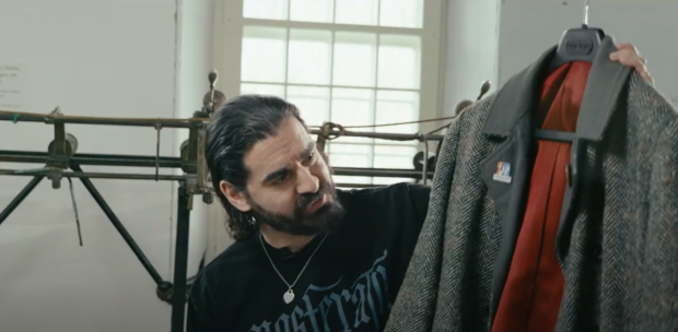 Close-up of someone sitting in front of a track sewing machine, holding up a peacoat on a hanger, in interview format.