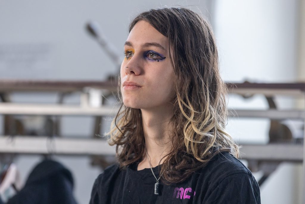 Portrait photo taken from the shoulders up. The person is angled to the left and looking straight ahead. She is wearing a black t-shirt with a small purple text on the right side reading MCR. She is wearing purple and yellow eye makeup.