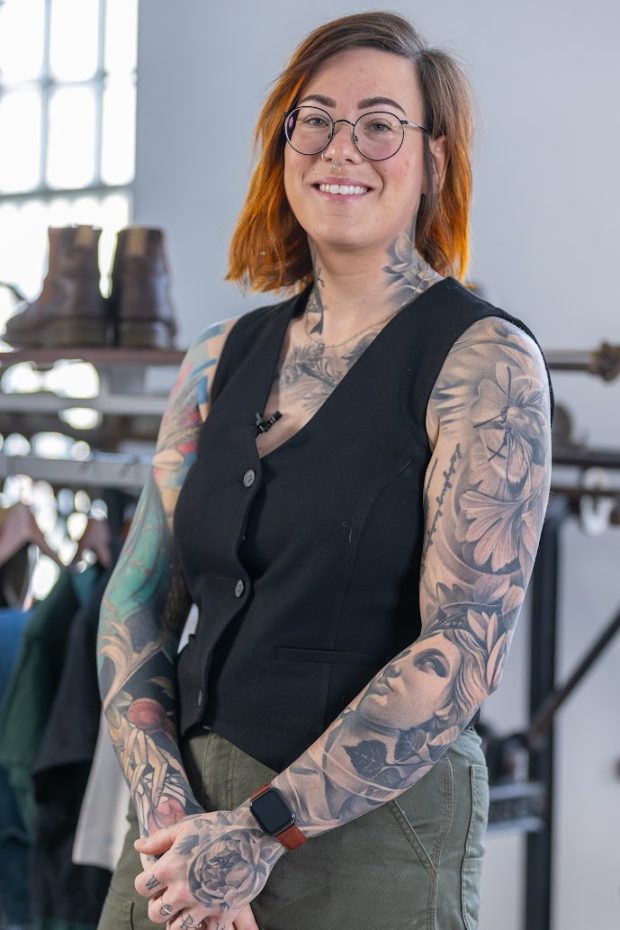 Portrait photo taken from the mid-thigh up. The person is smiling, wearing round, black-framed glasses and looking at the camera. She is wearing green pants and a black button-down vest. She has tattoos covering her arms and chest, going up the sides of her neck.