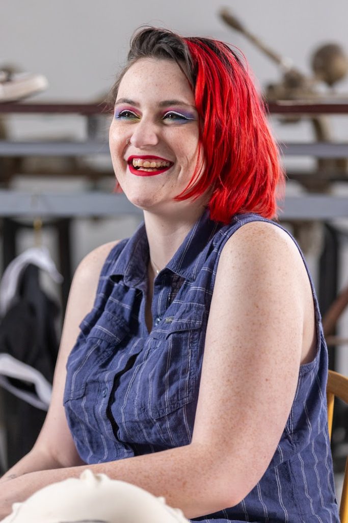 Portrait en plan taille. La personne est inclinée vers la gauche et regarde droit devant elle en riant. Iel a les cheveux rouge vif et le maquillage des yeux est violet, rose et blanc. Iel porte une chemise boutonnée, sans manches, bleu foncé à fines rayures blanches. 