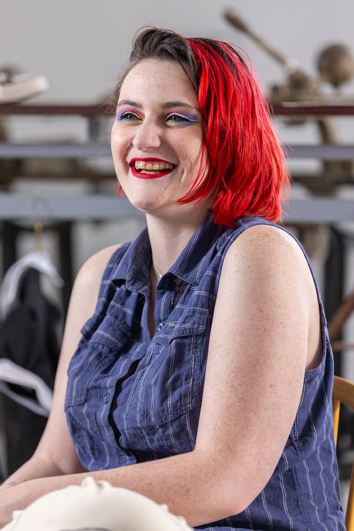 Portrait photo taken from the waist up. The person is angled towards the left and looking straight ahead, laughing. They have bright red hair and purple, pink, and white eye makeup. They are wearing a sleeveless button-down shirt that is dark blue with a white pinstripe.