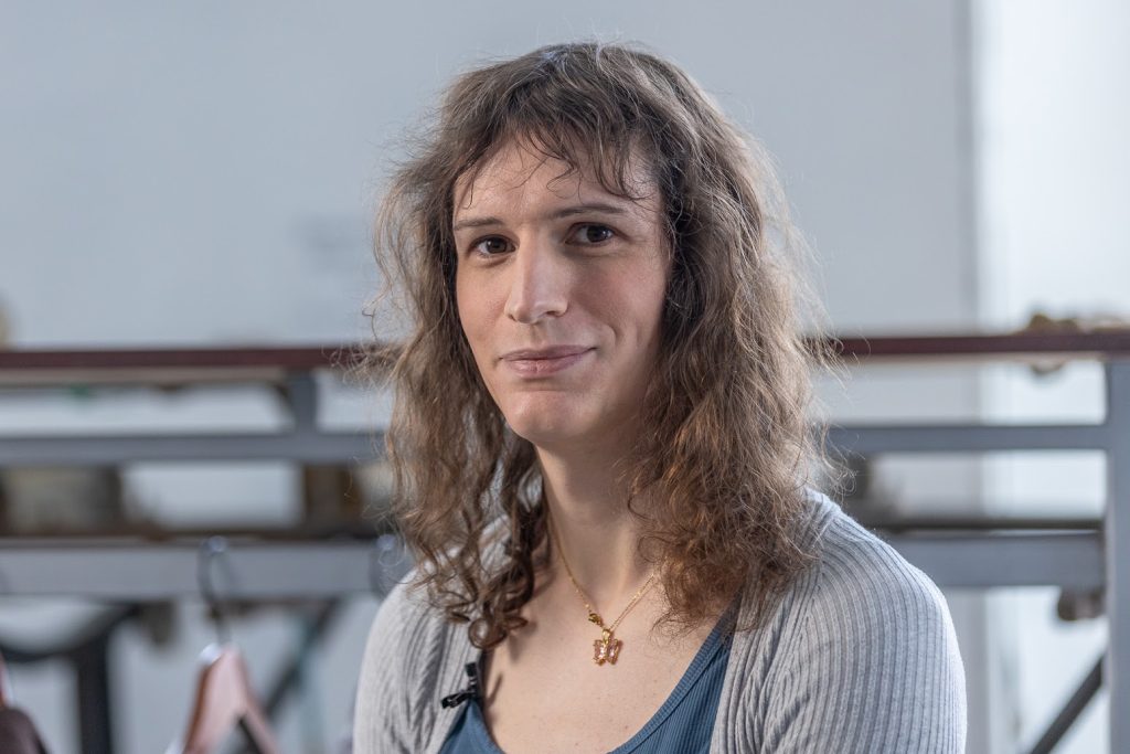 Portrait photo taken from the chest up. The person is smiling and looking at the camera. She is wearing a ribbed grey cardigan and a dark blue, scoop-necked shirt underneath, and a gold necklace.