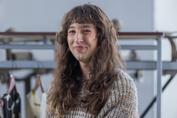 Portrait photo taken from the chest up. The person is smiling and looking at the camera. She is wearing a knit grey, brown, and orange sweater.