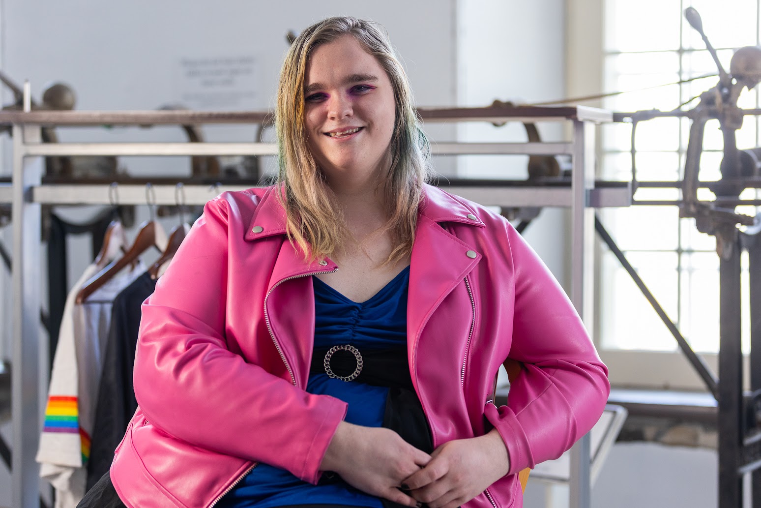 Portrait photo taken from the hips up. The person is smiling and looking at the camera. She is wearing a pink faux-leather jacket over a dark blue and black shirt with a silver buckle. She has pink eye makeup.