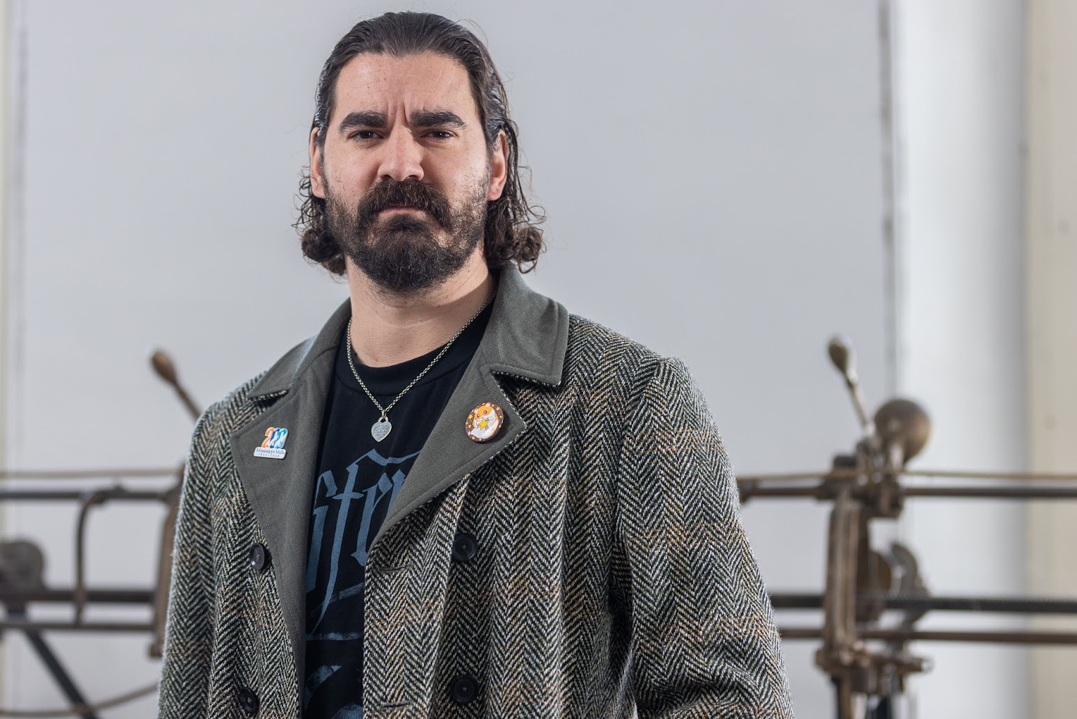 Portrait photo taken from the waist up. The bearded person is looking seriously into the camera. He is wearing a grey and black peacoat with a pin on either lapel. Underneath, he is wearing a black shirt with blue lettering.