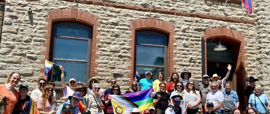 A group of people are gathered outside a stone building, lined up to take a group photo. A Pride flag is held up in the middle of the crowd, and another flies on a flagpole above the building's entrance. People hold smaller Pride flags of their own. They are smiling at the camera.
