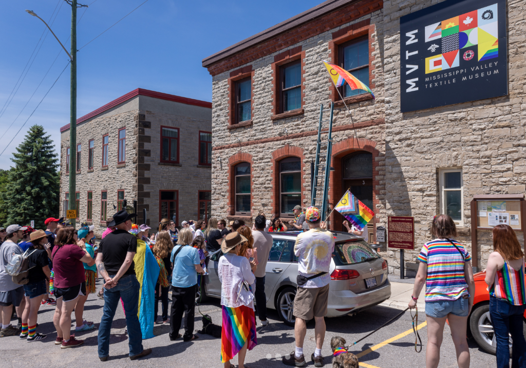 A crowd of people gathered in front of a stone building. A ladder extends up to a flag pole above the building's entrance, flying a Pride flag.