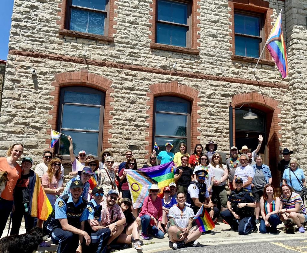 A group of people are gathered outside a stone building, lined up to take a group photo. A Pride flag is held up in the middle of the crowd, and another flies on a flagpole above the building's entrance. People hold smaller Pride flags of their own. They are smiling at the camera.