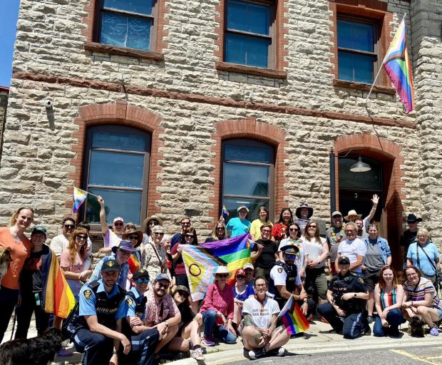 A group of people are gathered outside a stone building, lined up to take a group photo. A Pride flag is held up in the middle of the crowd, and another flies on a flagpole above the building's entrance. People hold smaller Pride flags of their own. They are smiling at the camera.