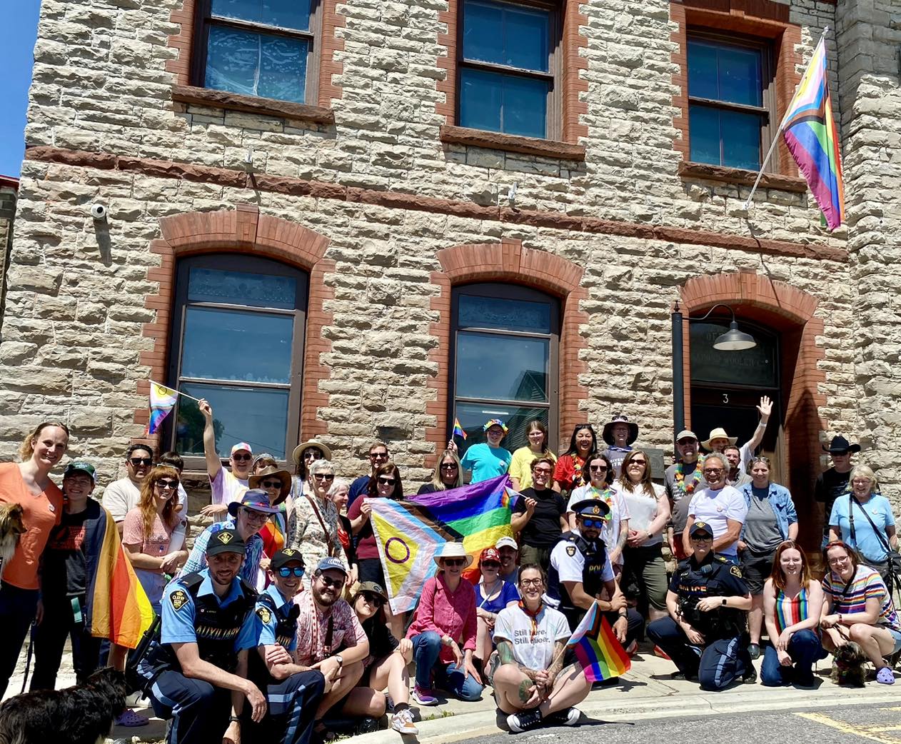 A group of people are gathered outside a stone building, lined up to take a group photo. A Pride flag is held up in the middle of the crowd, and another flies on a flagpole above the building's entrance. People hold smaller Pride flags of their own. They are smiling at the camera.