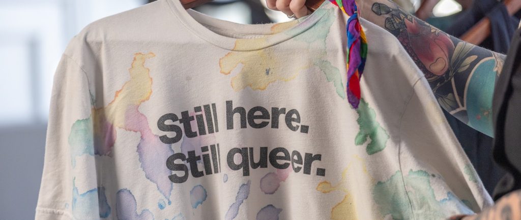 A tattooed arm is holding up an off-white t-shirt on a wooden hanger. The shirt has a multicoloured pattern of random, amorphous blobs resembling ink or watercolour. Black text across the front of the shirt reads Still here. Still queer. A rainbow-striped handkerchief is tied around the top of the hanger.