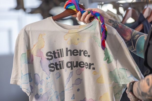 A tattooed arm is holding up an off-white t-shirt on a wooden hanger. The shirt has a multicoloured pattern of random, amorphous blobs resembling ink or watercolour. Black text across the front of the shirt reads Still here. Still queer. A rainbow-striped handkerchief is tied around the top of the hanger.
