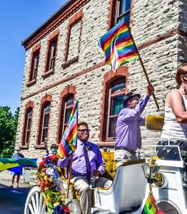 2 people wearing lavender shirts on a white carriage, waving Pride flags. Another person is cut out of the frame, at the head of the carriage. They are in front of a stone building.