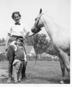 A black and white photograph of a woman, young boy and a horse standing in a park; several cars, buildings and people are in the background behind a temporary fence