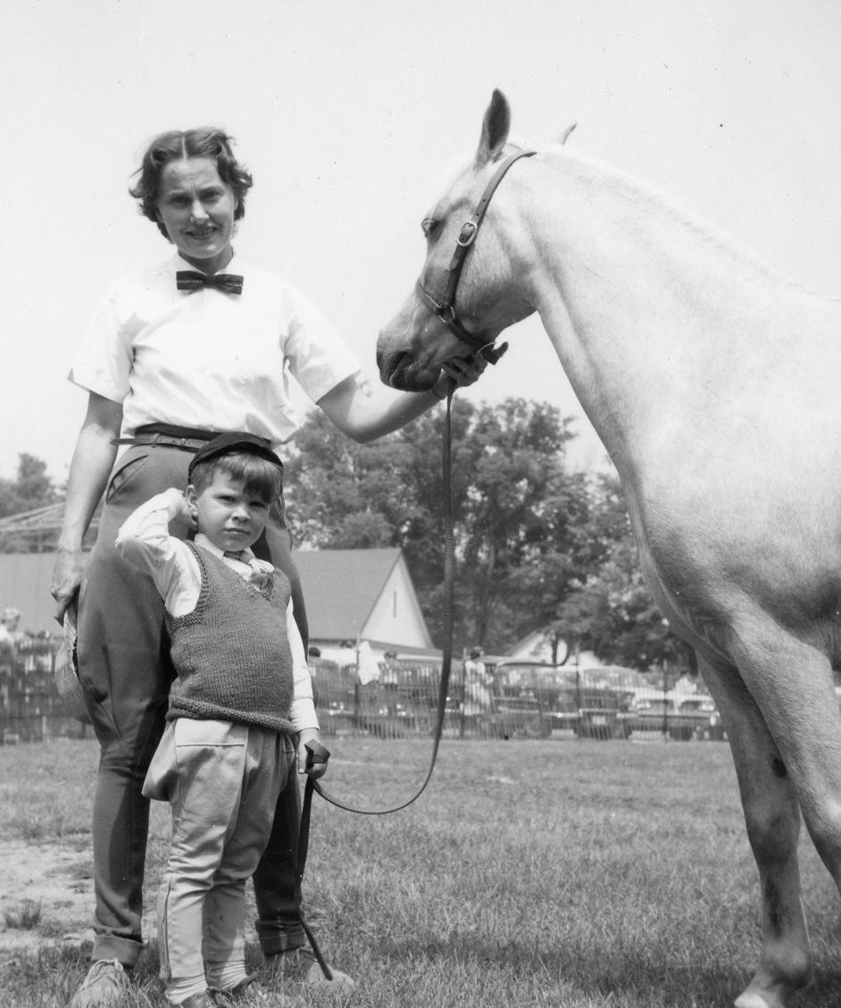 A black and white photograph of a woman, young boy and a horse standing in a park; several cars, buildings and people are in the background behind a temporary fence