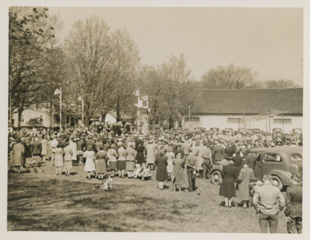 A somewhat blurry black and white photograph of a large gathering of people, most with their backs facing the camera, in a park surrounding a raised platform at the left side with four posts bearing Union Jack flags, upon which are seated about a dozen people; large white clad building and several trees and houses in the background
