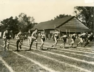 Photo en noir et blanc de neuf garçons en short et débardeur courant sur des pistes dans un parc.  Grand bâtiment en bois et quelques spectateurs et voitures à l’arrière.