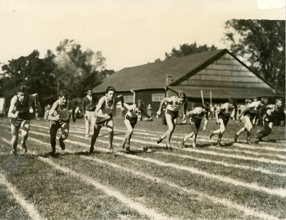 A black and white photograph of group of nine boys wearing shorts and tank tops running in lanes painted on grass in a park setting; large rectangular wooden building with a few spectators and cars in the background