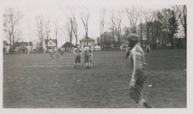 A black and white photograph of a group of boys in football uniforms spread out across a grass field; houses are visible in the background behind a picket fence and a row of trees