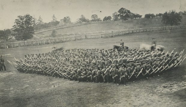 A black and white photograph of soldiers in a field with rifles drawn in a tight, square formation, with three horses, two are partially blurred, at the back; two men look on from the left; a farm and wooden fencing is visible in the background