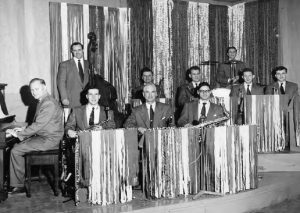 A somewhat blurry black and white photograph of a ten piece male band wearing dress suits and ties, sitting behind covered music stands, on two levels on a raised double platform; the music stands and background covered by panels of paper or fabric vertical strips giving a shimmering effect; piano and player at bottom left