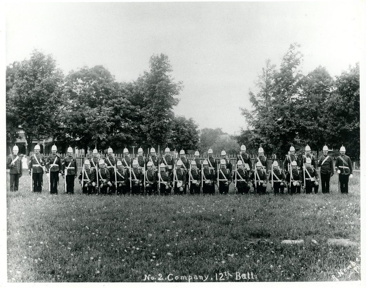 A black and white photograph of male soldiers posing in a park setting, in two rows, front row on right knee; all holding a rifle or sword and wear uniforms with helmet containing a spike on top; wooden picket fence, trees and obscured houses in background