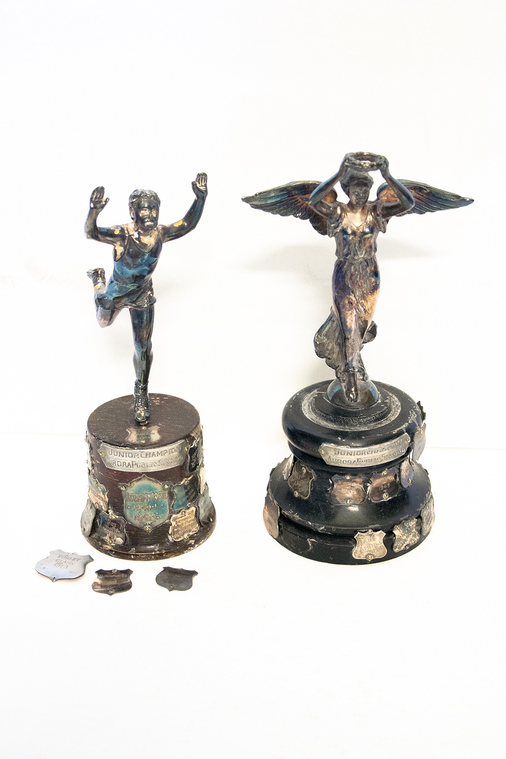 Two small silver trophies on wooden bases covered with engraved silver shields; the one at left depicts a male figure running with both arms raised; the one at right features a winged female figure in flowing dress with raised arms holding a wreath with both hands; three loose silver engraved shields sit in front of the trophy at left