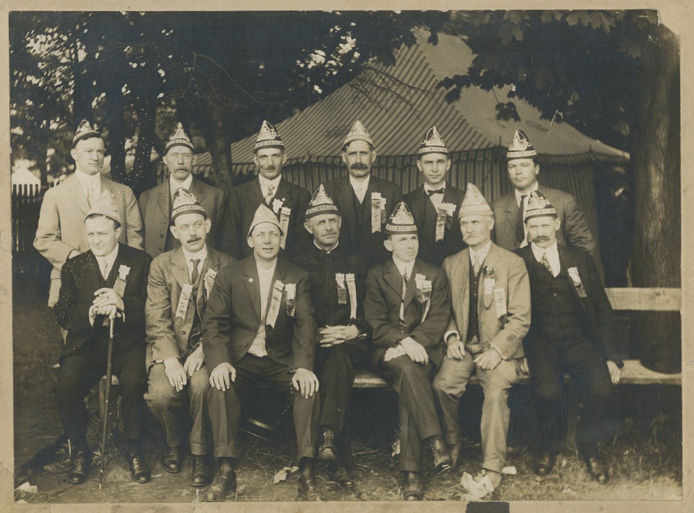 A black and white photograph of a group of 13 men in suits wearing the same triangular shaped hats and matching pins and ribbons pose in two rows, front row seated on bench, in front of a striped marquee in a treed setting