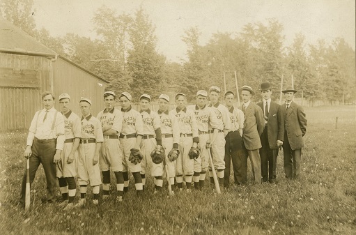 A black and white photograph of fourteen men, some wearing baseball gloves and others holding bats, posing in a straight line on a grass field with trees and a wooden building in the background; ten men are wearing baseball uniforms inscribed with Best Everyday Shoe on front