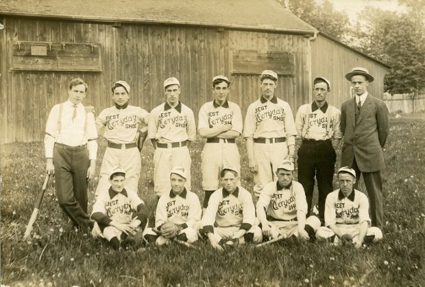 A black and white photo of a male baseball team posing in two rows, with the front row seated on the grass, in front of a wooden building; 10 men wear uniforms with the BEST Everyday SHOE written on front, two men stand at either end of back row in dress clothing