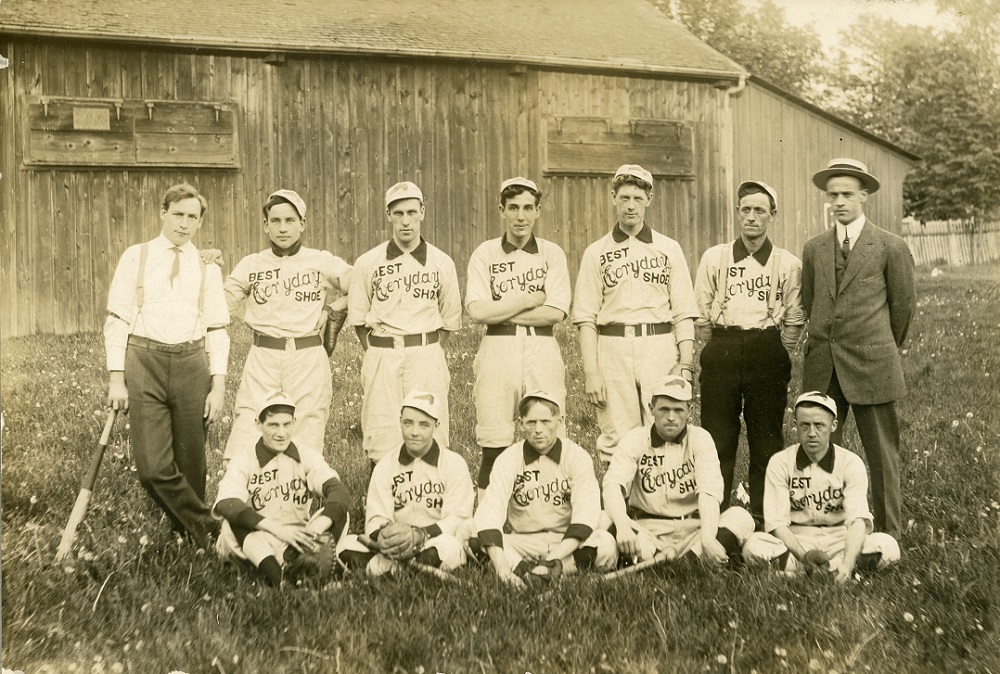 A black and white photo of a male baseball team posing in two rows, with the front row seated on the grass, in front of a wooden building; 10 men wear uniforms with the "BEST Everyday SHOE" written on front, two men stand at either end of back row in dress clothing